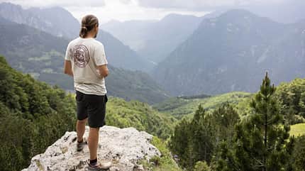 Etappe 3, Theth, Valbonё, Valbone Pass, Albanien, Wanderung, Gebirge, Berglandschaft, Anstieg, Outdoor Eine Person steht auf einem Felsen und blickt in ein bewaldetes Gebirgstal mit Bergen im Hintergrund. Die Szene zeigt eine Wanderetappe in Albanien zwischen Theth und Valbonё am Valbone Pass.