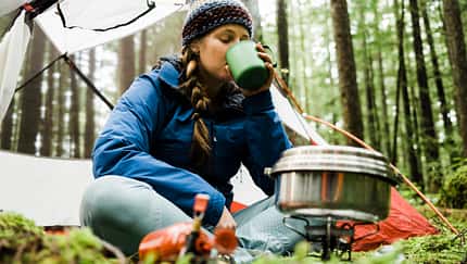 GettyImages/Alex Ratson: A blonde haired young women sits in front of her camping tent in a lush rainforest with a camp stove and pot steaming in front of her drinks a warm beverage from a mug